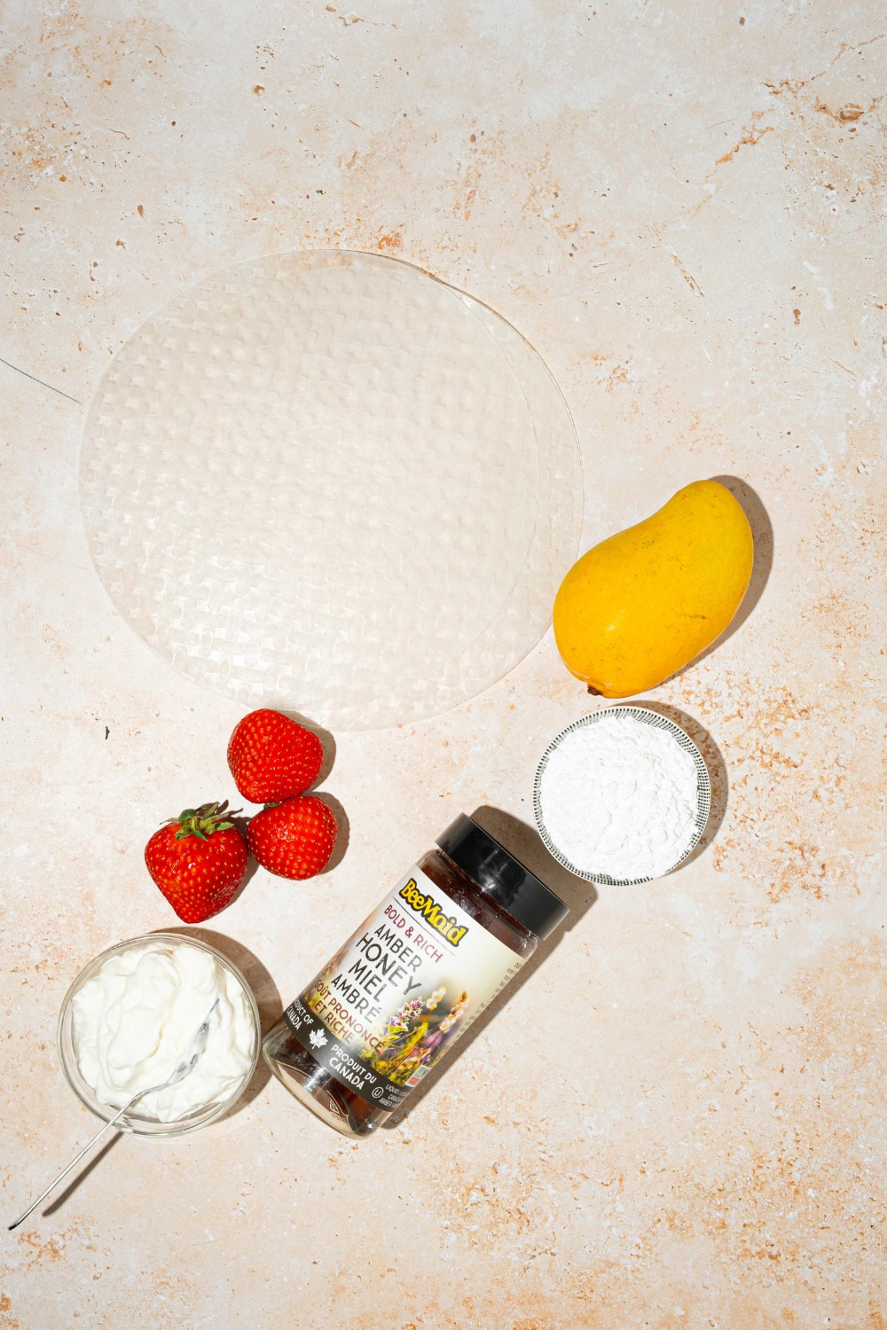A tan counter with ingredients to make rice paper mochi including rice paper sheets, mango, strawberries, honey, yogurt, and rice flour.