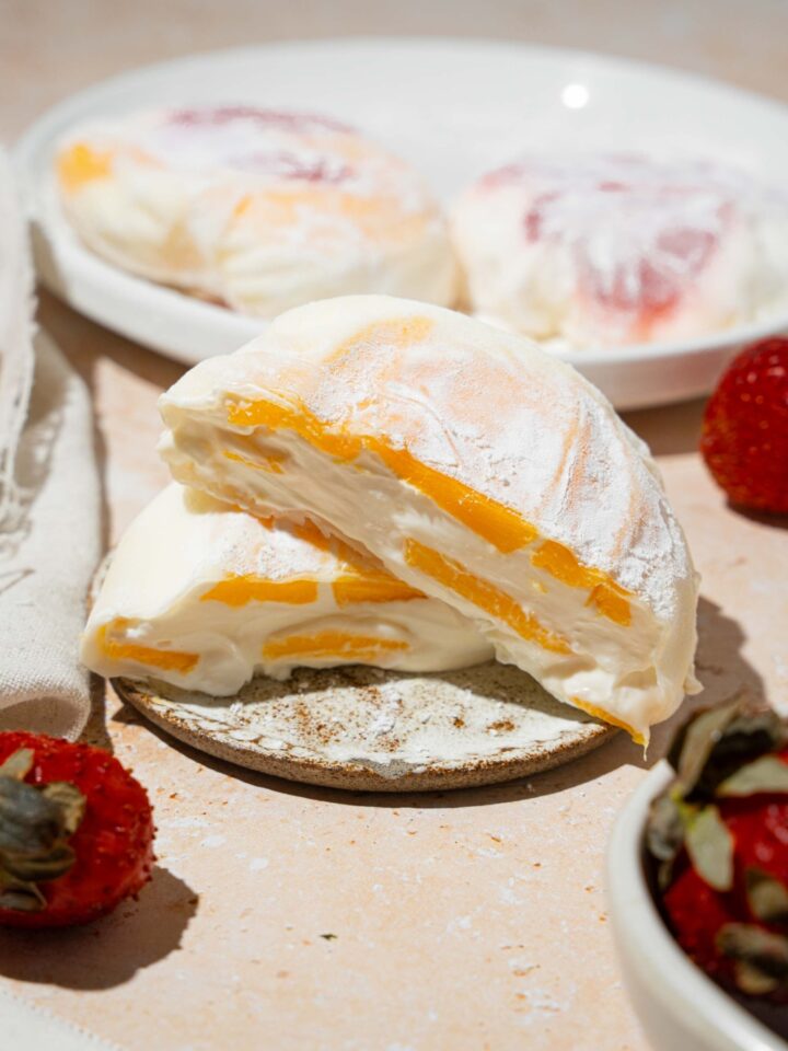 A plate with mango rice paper mochi coated in rice flour and sliced in half. The plate is on a tan counter with an additional plate of mochi and bowl of strawberries.