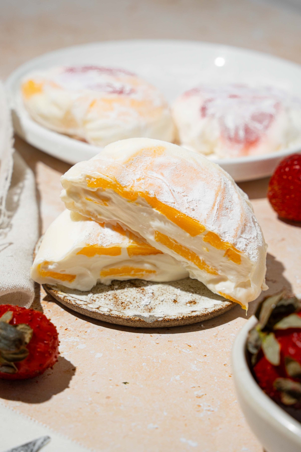 A plate with mango rice paper mochi coated in rice flour and sliced in half. The plate is on a tan counter with an additional plate of mochi and bowl of strawberries.