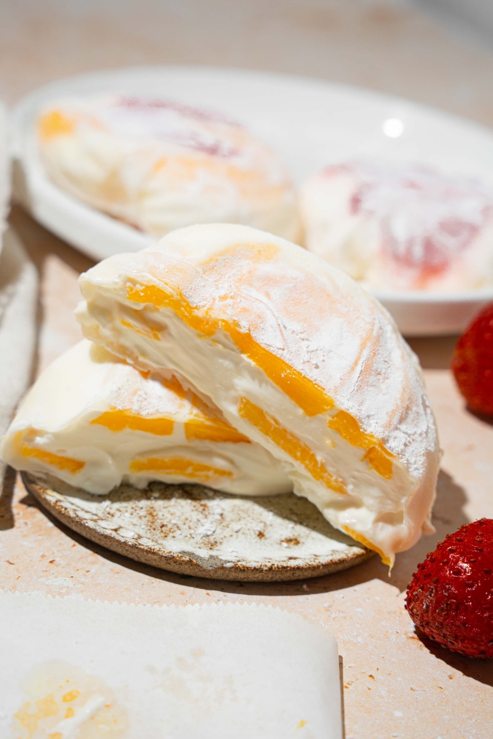 A plate with mango rice paper mochi coated in rice flour and sliced in half. The plate is on a tan counter with an additional plate of mochi and strawberries.