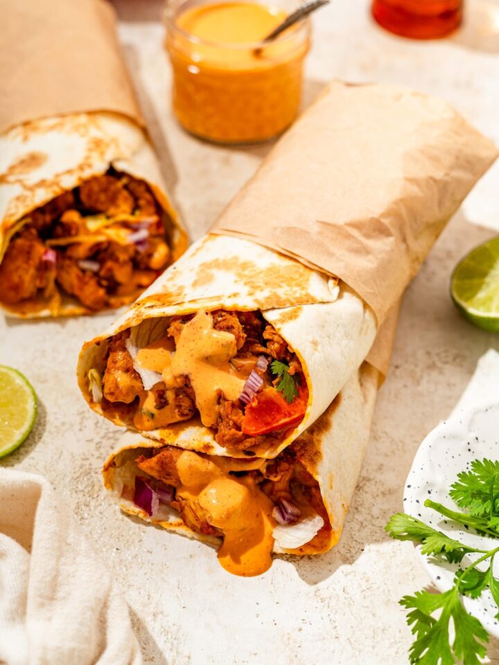 Two honey chipotle chicken wraps wrapped in paper and stacked on one another. The wraps are on a tan counter with an additional wrap and a bowl of dipping sauce.