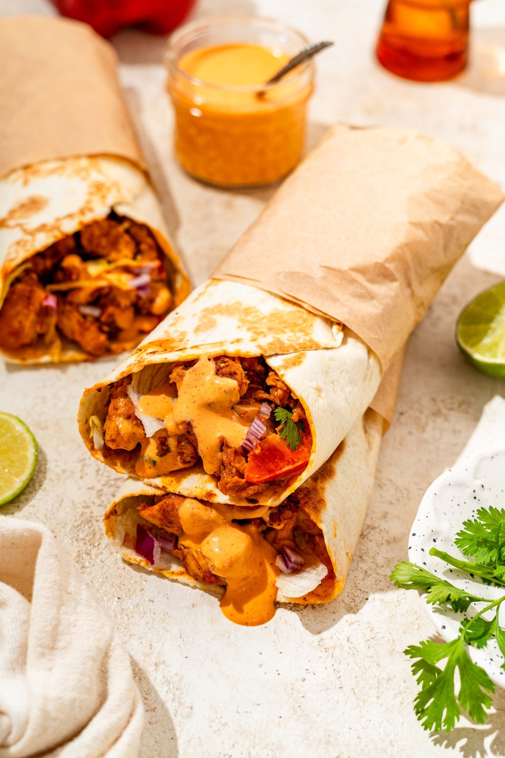 Two honey chipotle chicken wraps wrapped in paper and stacked on one another. The wraps are on a tan counter with an additional wrap and a bowl of dipping sauce.