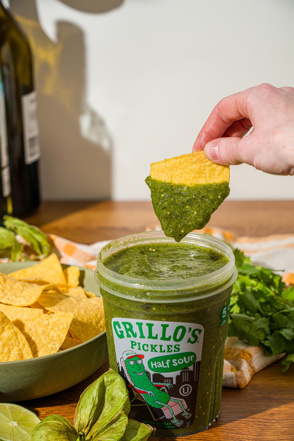 A chip dipping into pickle salsa in a Grillo's pickles container. The container is on a wooden counter with a bowl of tortilla chips.