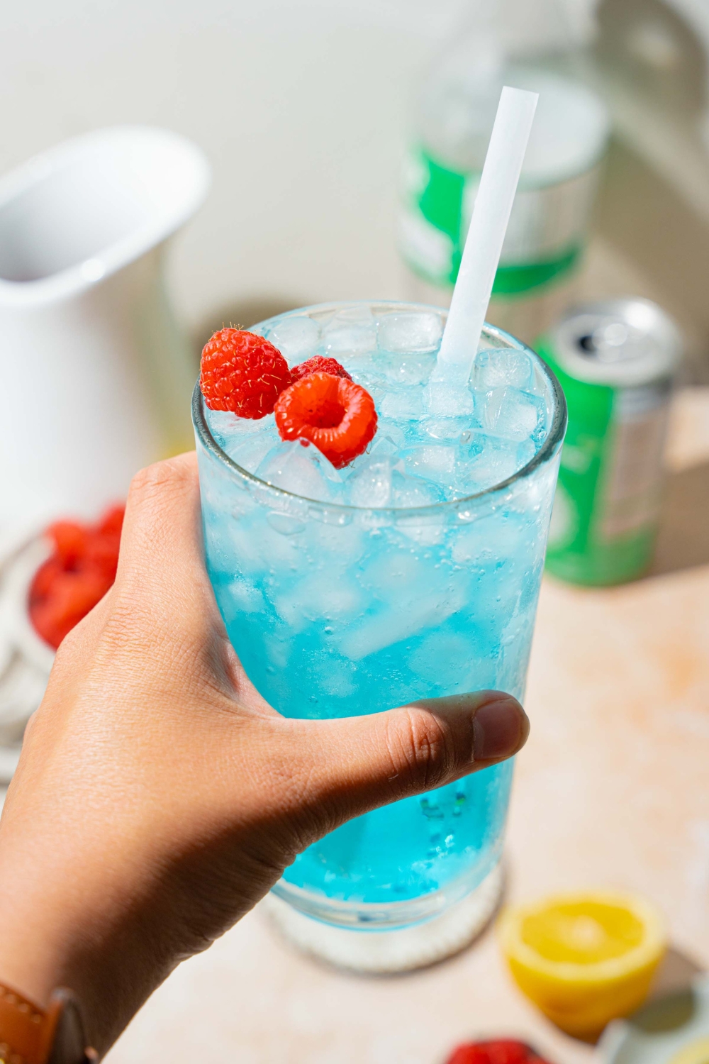 A close up of a hand holding a tall glass of ocean water drink served with pebble ice, a straw, and garnished with fresh raspberries. There is a tan counter with sliced lemon and a can of soda blurred in the background.