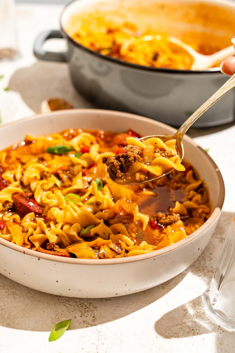 A white bowl with dirty cabbage and noodles with a spoon taking a bite from the bowl. The bowl is on a tan counter with a dutch oven of cabbage noodles.