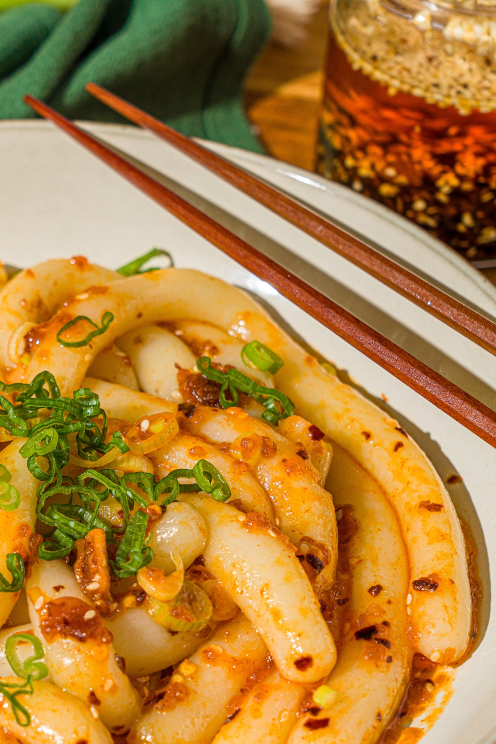 A white bowl with spicy potato noodles tossed in chili crisp and garnished with sliced green onion. A pair of chopsticks are resting on the bowl. The bowl is on a wooden counter with a green cloth napkin.