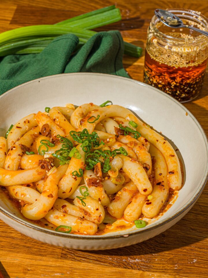 A white bowl with spicy potato noodles tossed in chili crisp and garnished with sliced green onion. The bowl is on a wooden counter with a green cloth napkin and jar of chili crisp.