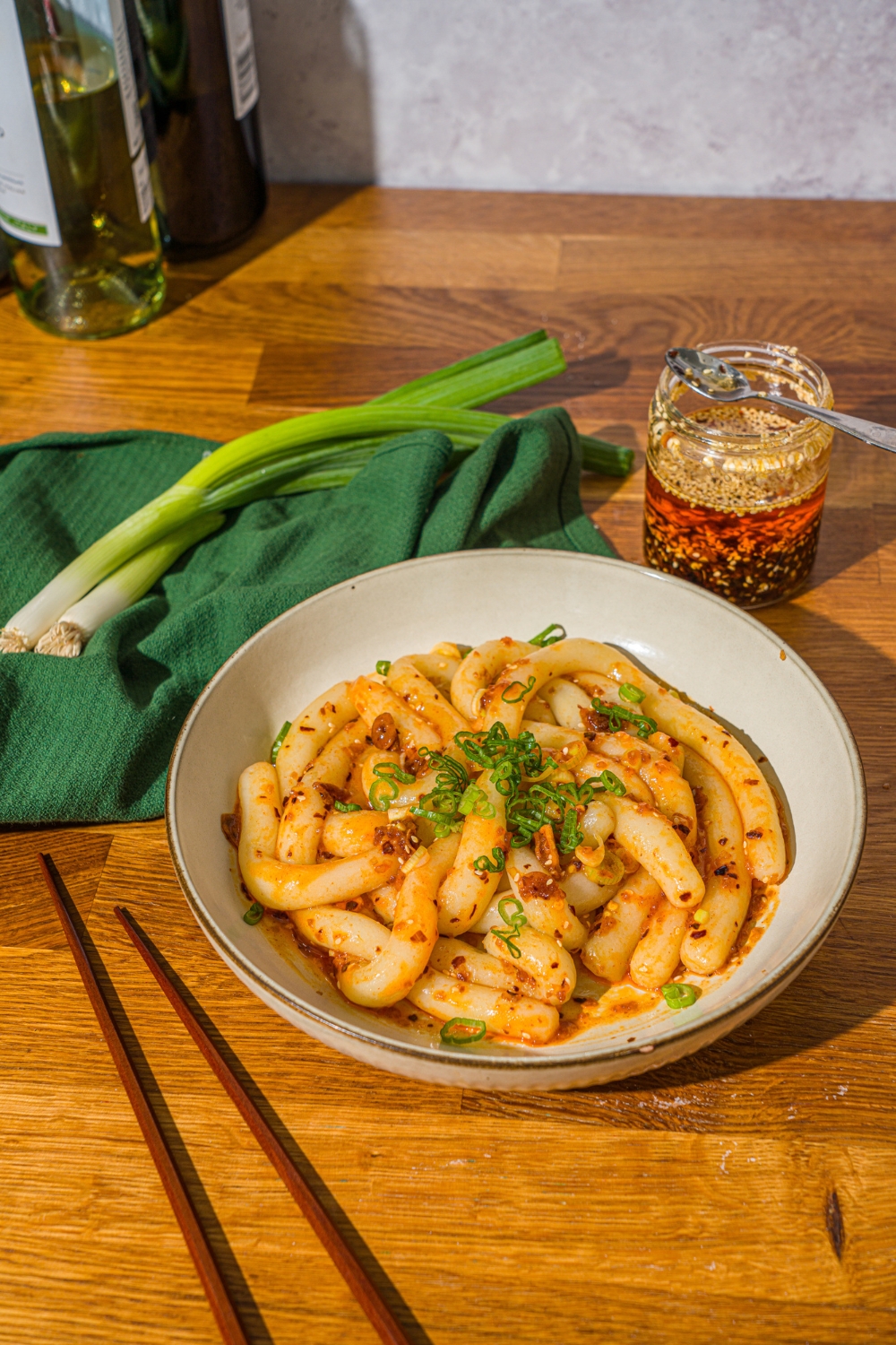 A white bowl with spicy potato noodles tossed in chili crisp and garnished with sliced green onion. The bowl is on a wooden counter with a pair of chopsticks, green cloth napkin, and jar of chili crisp.