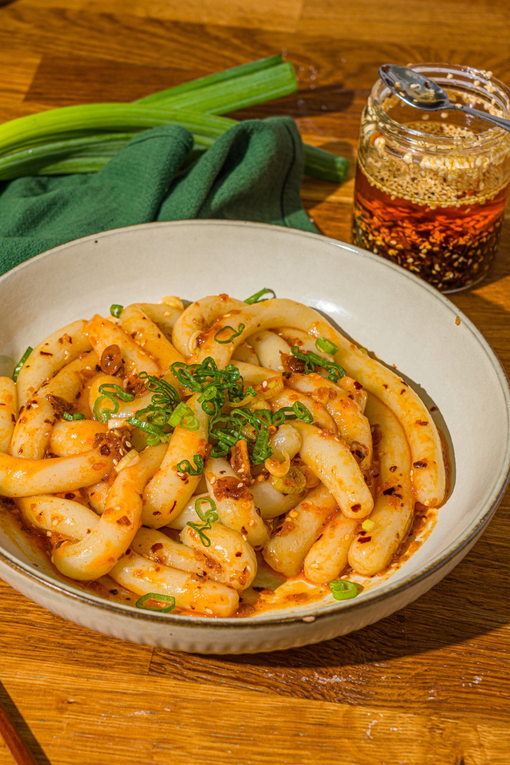 A white bowl with spicy potato noodles tossed in chili crisp and garnished with sliced green onion. The bowl is on a wooden counter with a green cloth napkin and jar of chili crisp.