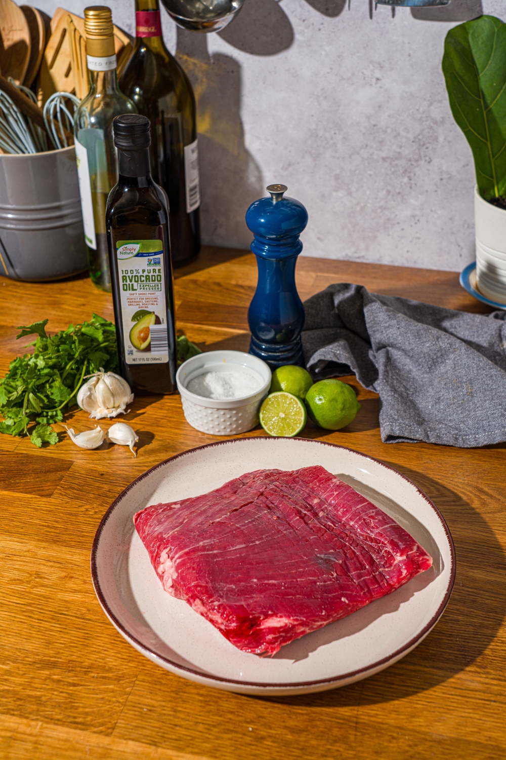 A wooden counter with ingredients to make steak bowls with cilantro lime sauce including steak, lime, cilantro, olive oil, garlic, salt, and pepper.