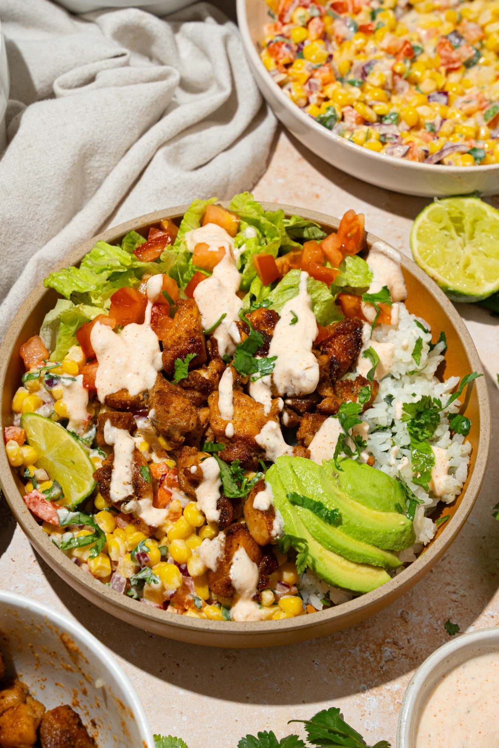 A brown bowl with street corn chicken rice bowl served with cilantro lime rice, seasoned chicken, street corn, and avocado, drizzled in a greek yogurt dressing. The bowl is on a tan counter with a bowl of street corn and a white cloth napkin.