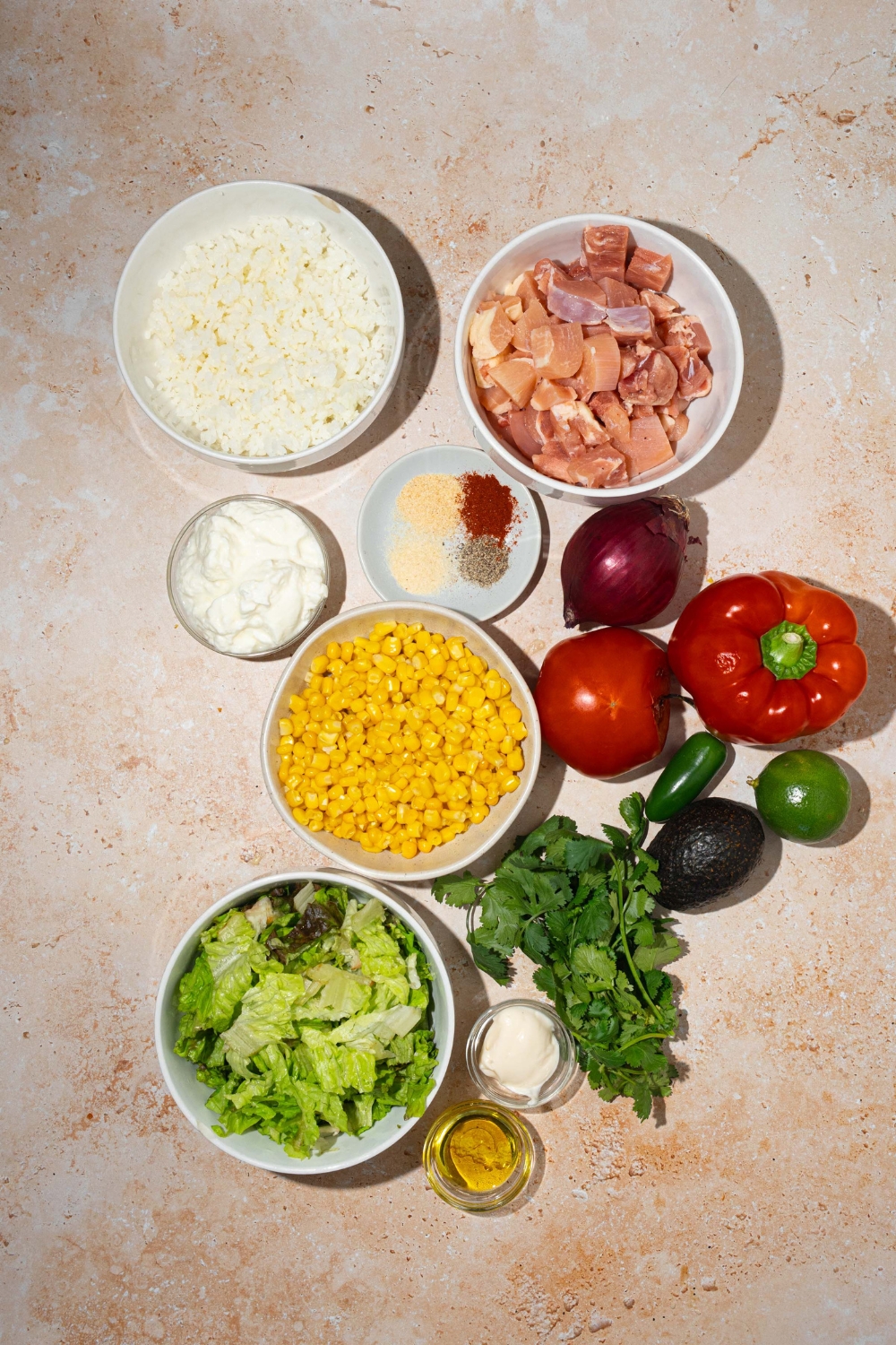 A tan counter with several bowls containing ingredients to make street corn chicken rice bowl including rice, chicken, corn, lettuce, red pepper, jalapeño, avocado, greek yogurt, mayo, oil, and seasonings.