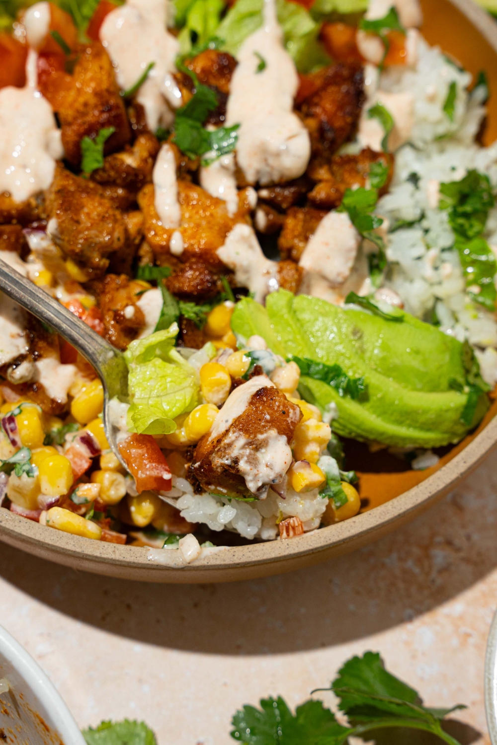 A brown bowl with street corn chicken rice bowl served with cilantro lime rice, avocado, street corn mixture, and seasoned chicken drizzled with greek yogurt dressing. There is a fork in rice bowl.