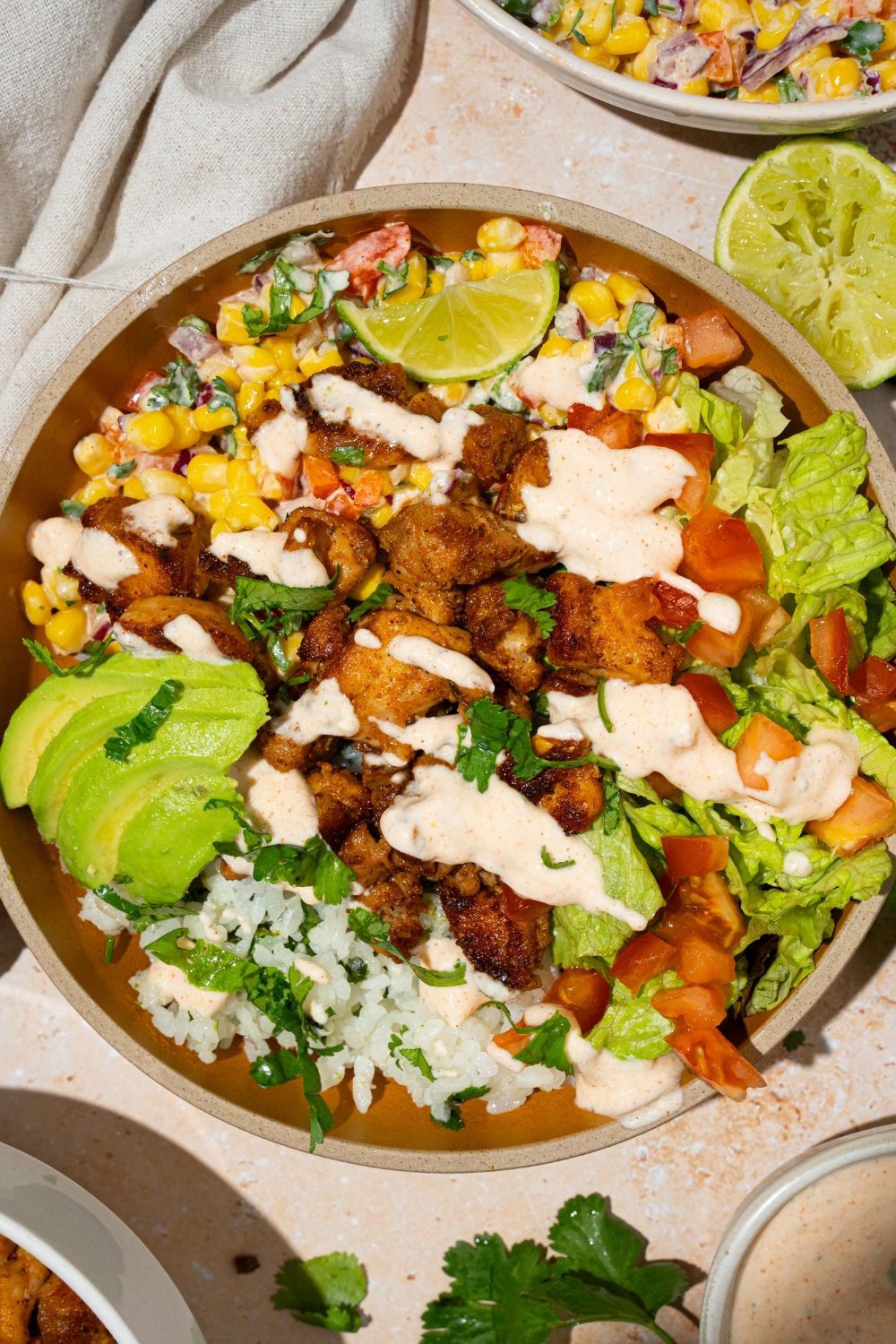 A brown bowl with street corn chicken rice bowl served with cilantro lime rice, seasoned chicken, street corn, and avocado, drizzled in a greek yogurt dressing. The bowl is on a tan counter with a bowl of street corn and a white cloth napkin.