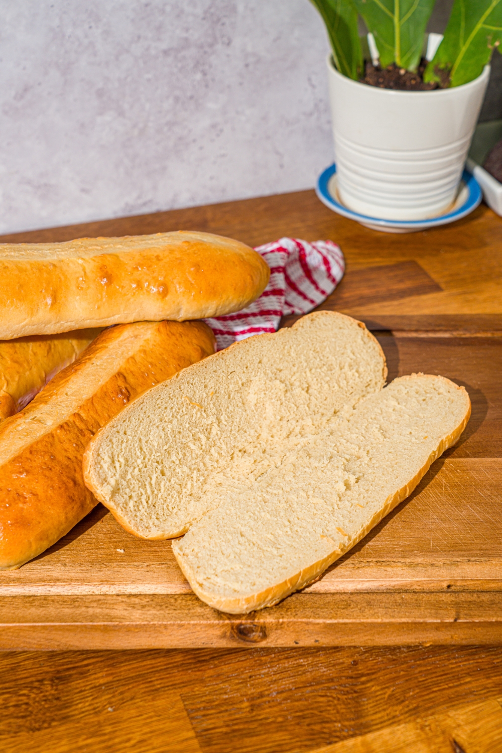 A wooden cutting board with homemade hoagie rolls. One of the rolls is sliced down the middle. The board is on a wooden counter with a red striped napkin.