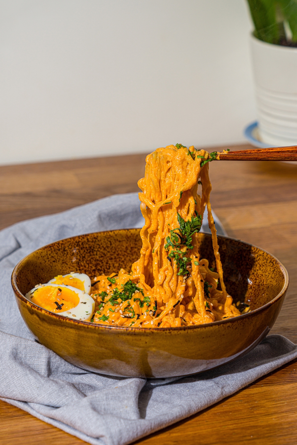 A bowl of Buldak ramen with soft boiled egg and garnished with fresh parsley and sesame seeds. A pair of chopsticks are pulling noodles from the bowl. The bowl is on a wooden counter with a blue cloth napkin.