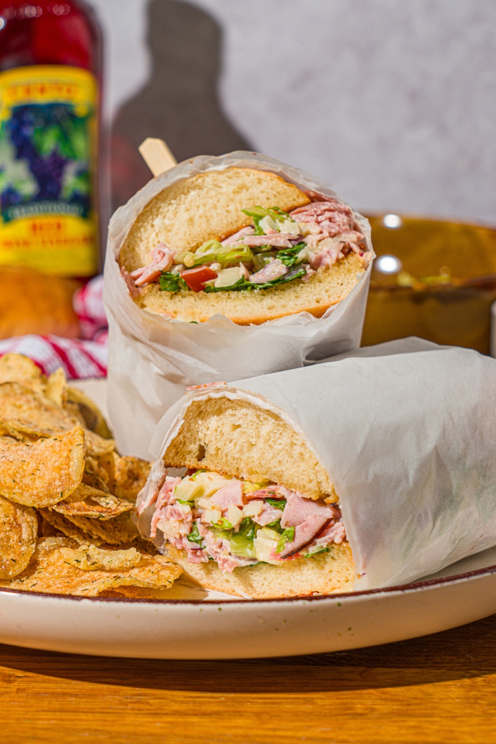 A white plate with a chopped Italian sandwich wrapped in paper and sliced in half, and chips. The plate is on a wooden counter.