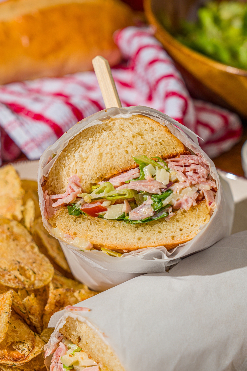 A white plate with a chopped Italian sandwich wrapped in paper and sliced in half, and chips. The plate is on a wooden counter with a bowl of salad and red striped napkin.