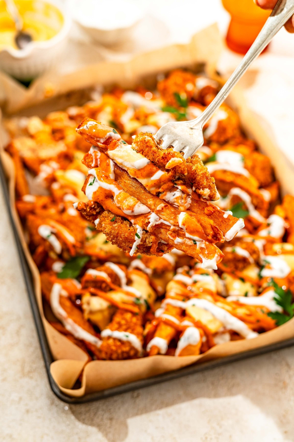 A close up of a fork with a bite of loaded fries with crispy chicken. There is a baking sheet lined with parchment paper with loaded fries topped with crispy chicken, cheese sauce, sriracha mayo sauce, and yogurt sauce blurred in the background.