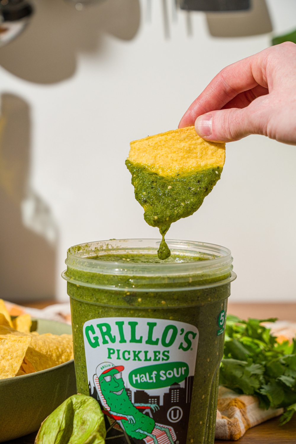 A chip dipping into pickle salsa in a Grillo's pickles container. The container is on a wooden counter with a bowl of tortilla chips.