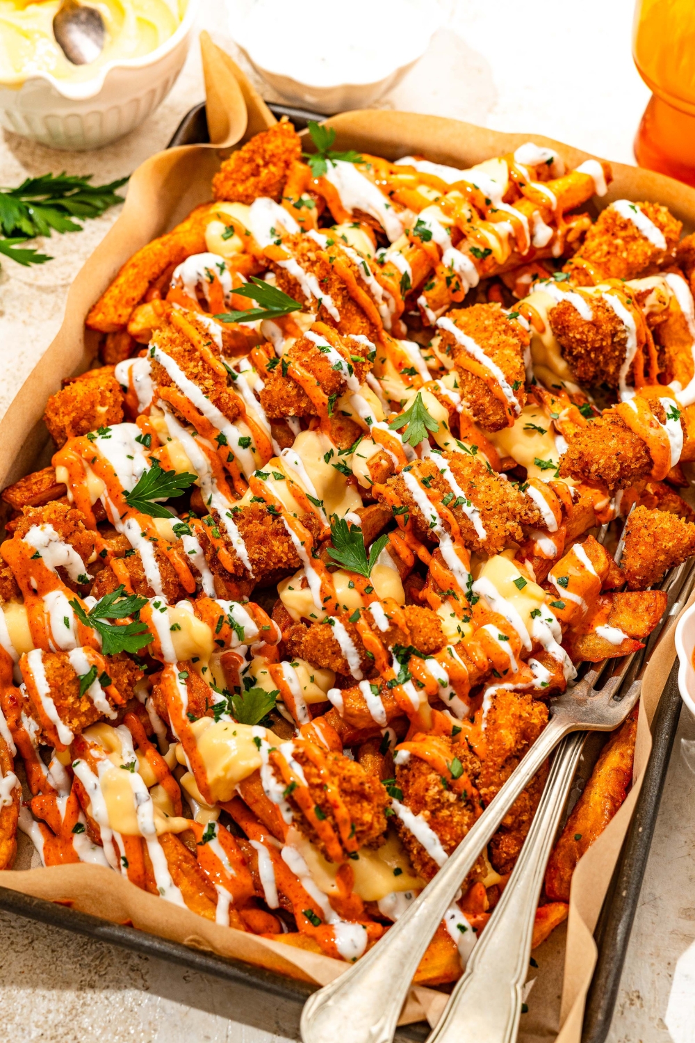 A baking sheet lined with parchment paper with loaded fries topped with crispy chicken, sriracha mayo sauce, yogurt sauce, cheese sauce, and fresh parsley. There are a pair of forks in the fries. The sheet is on a tan counter with bowls of toppings.