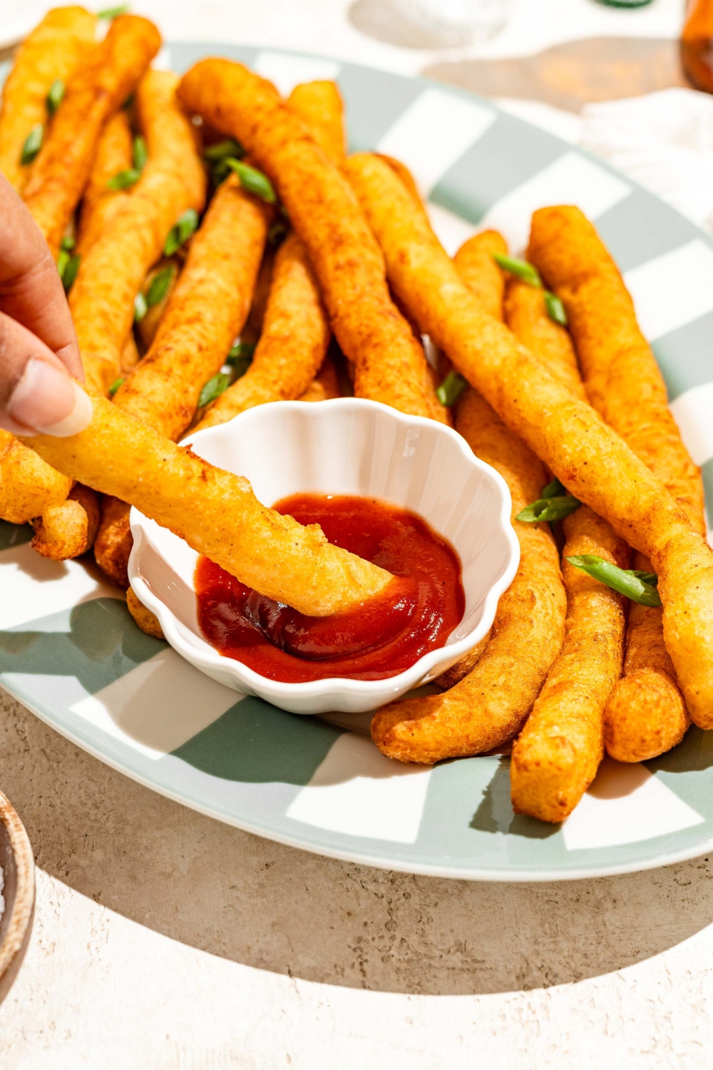 A striped plate with fried Japanese long fries garnished with sliced green onion and served with a small bowl of ketchup. A fry is being dipped into the ketchup. The plate is on a tan counter.