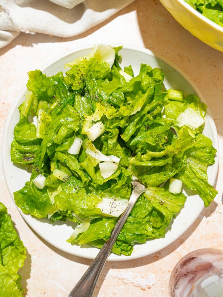 A white plate with wilted lettuce salad tossed in a bacon vinegar dressing. There is a fork in the salad. The plate is on a tan counter with a bowl of salad and white cloth napkin.