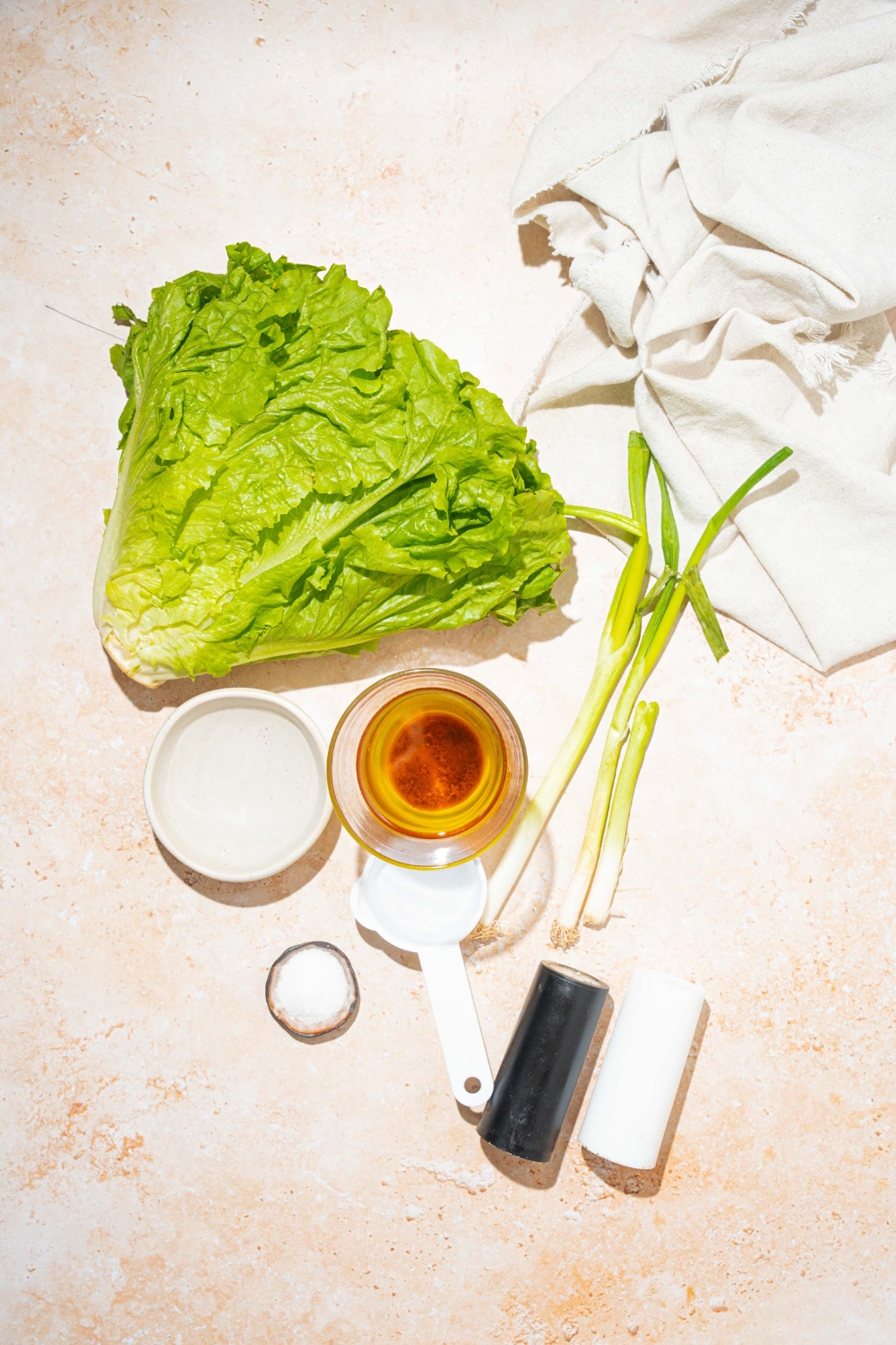 A tan counter with ingredients to make wilted lettuce salad including lettuce, bacon grease, green onion, vinegar, sugar, salt, and pepper.