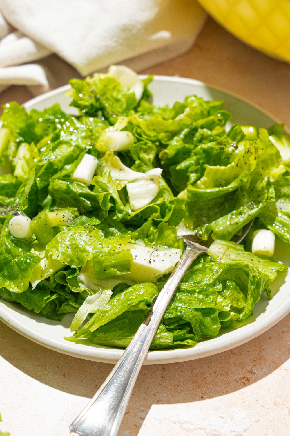 A white plate with wilted lettuce salad tossed in a bacon vinegar dressing. There is a fork in the salad. The plate is on a tan counter with a white cloth napkin.