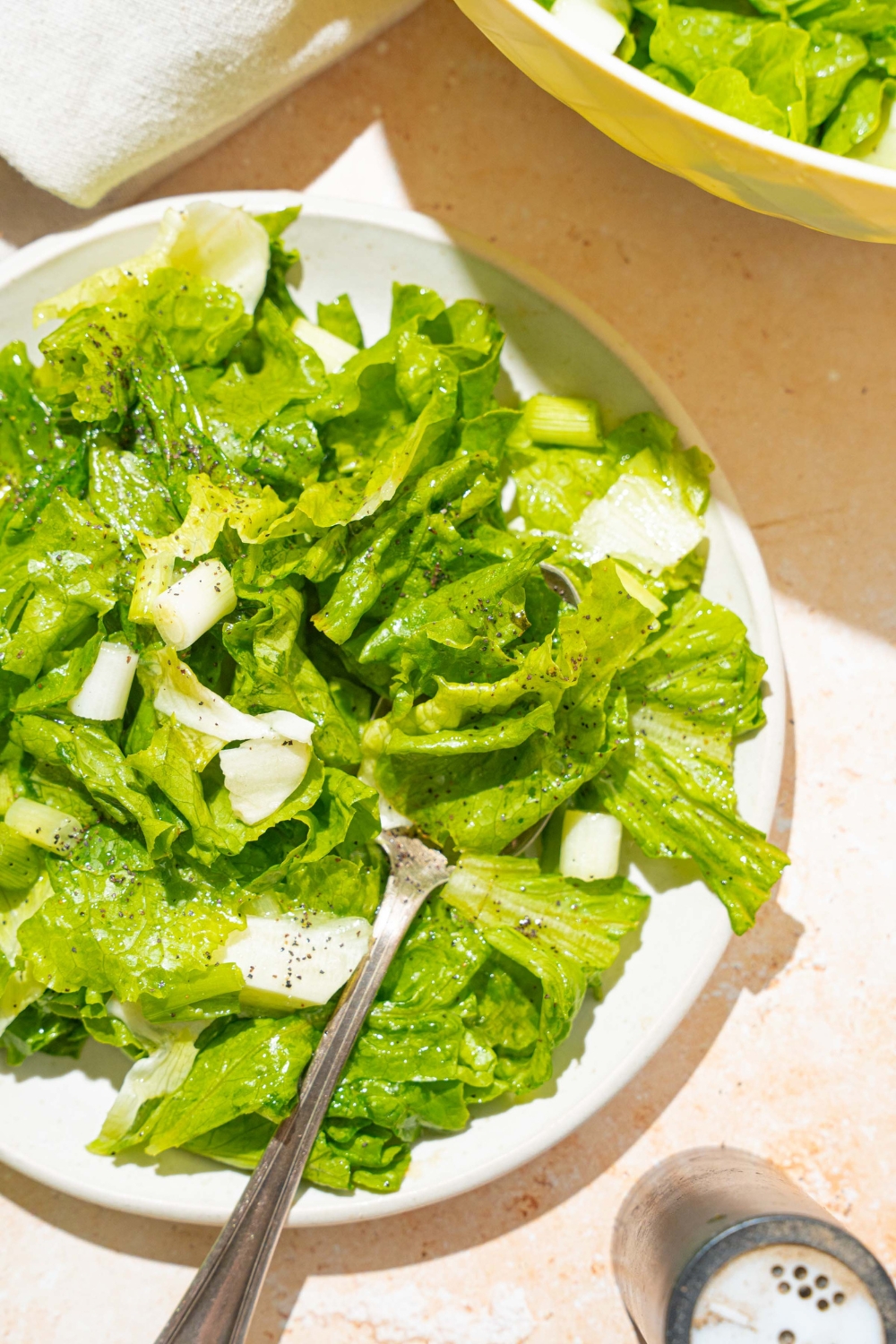 A white plate with wilted lettuce salad tossed in a bacon vinegar dressing. There is a fork in the salad. The plate is on a tan counter with a bowl of salad and white cloth napkin.