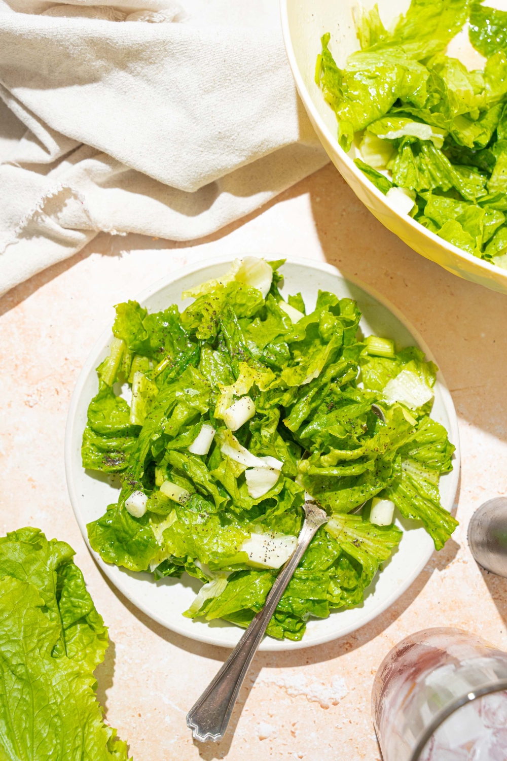 A white plate with wilted lettuce salad tossed in a bacon vinegar dressing. There is a fork in the salad. The plate is on a tan counter with a bowl of salad and white cloth napkin.
