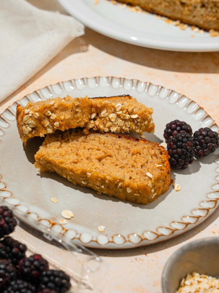 Two slices of rice cooker, banana bread with one leaning on the edge of the other and a few blackberries on a white plate.