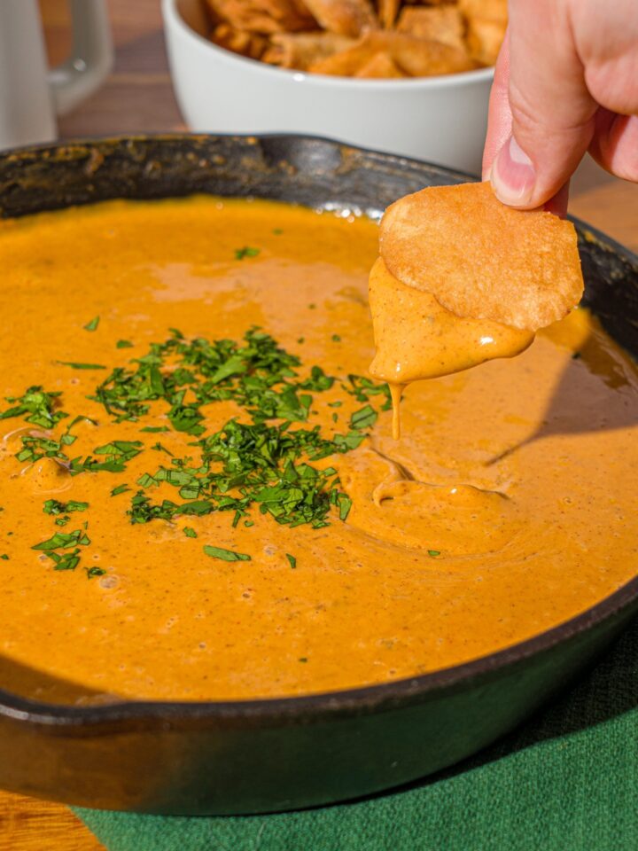A hand holding a chip with chili's skillet queso on it being held above a skillet filled with the queso.