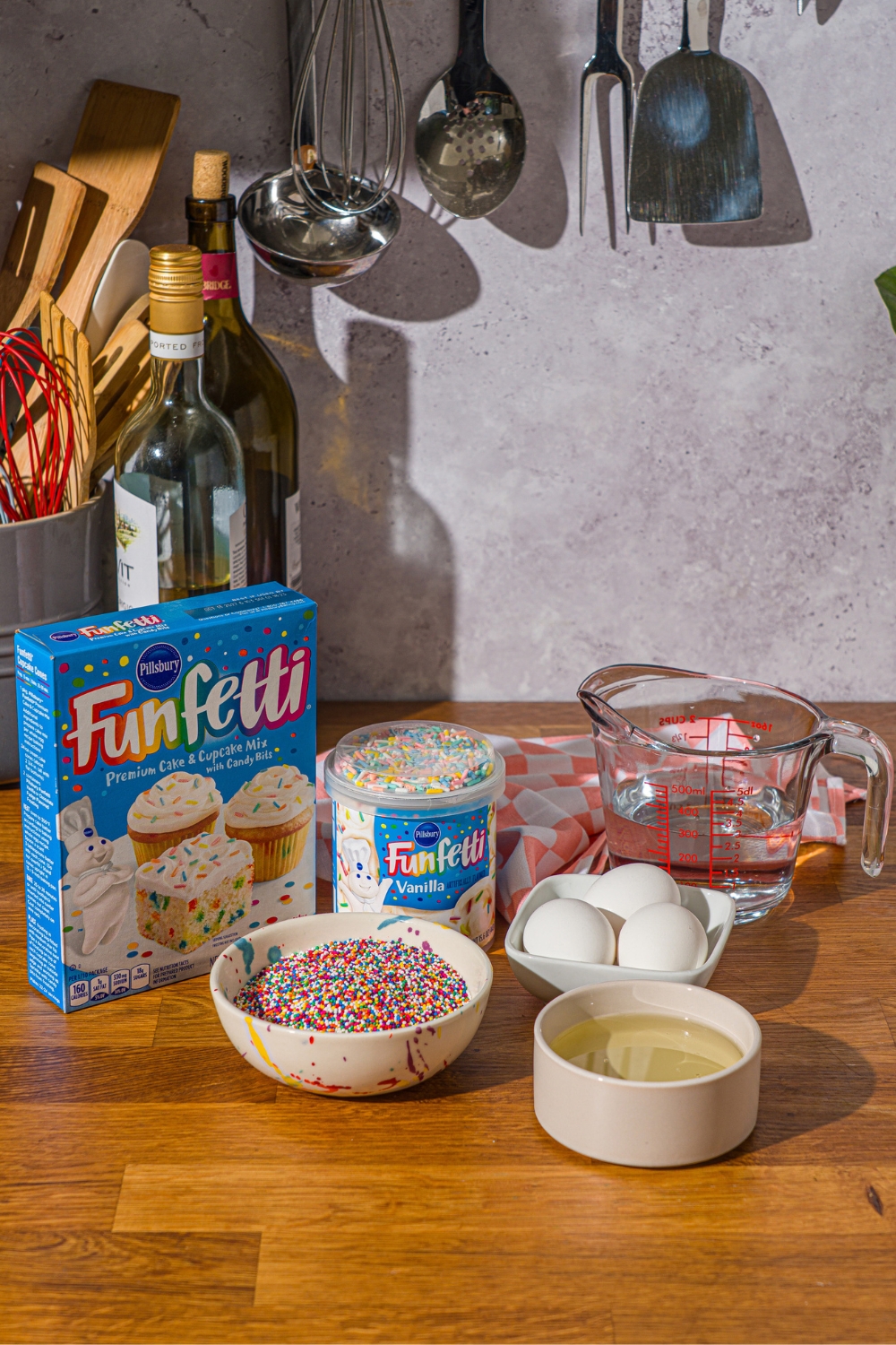 A box of funfetti, cake mix, a tub of funfetti frosting, a bowl with rainbow, pearl sprinkles, a bowl with three eggs in it, and a ptcher with water in it all on a wood countertop.