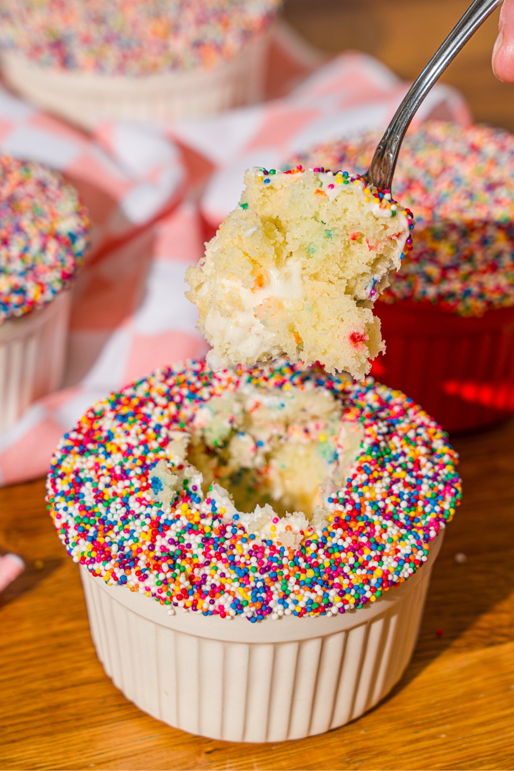 A spoon that has the inside of a dot cake on it being held over a dot cake that has rainbow sprinkles on top and is in a white ram.