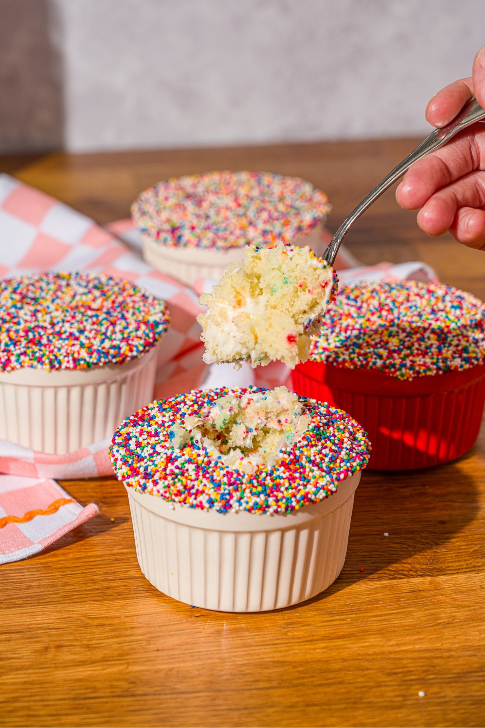 A hand holding a spoon that has funfetti cake, filling and vanilla frosting on it being held over a cake with rainbow sprinkles on top and a white ram.