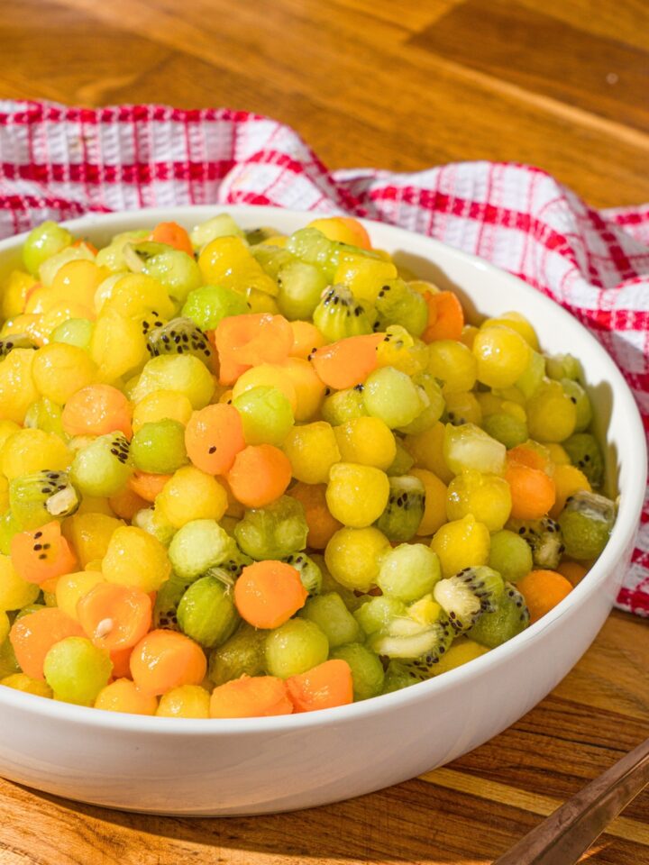 A dot fruit salad in a white. The fruit salad is small balls of yellow watermelon, honey dew, kiwi, and apricots.