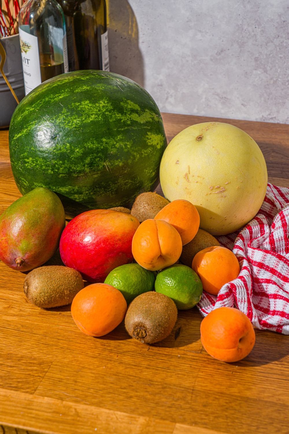 A watermelon, mangos, kiwis, apricots, and a honeydew melon all on a wooden counter.