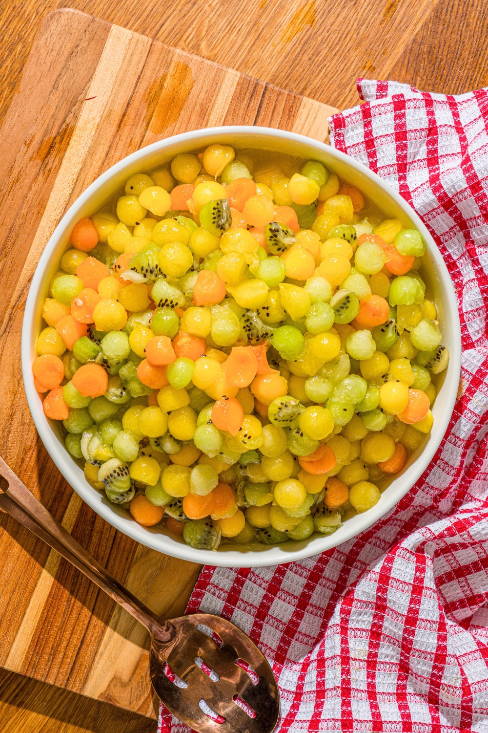 A dot fruit salad in a large white bowl on a wooden counter consisting of small spheres of honey dew, yellow watermelon, apricots, and kiwi.