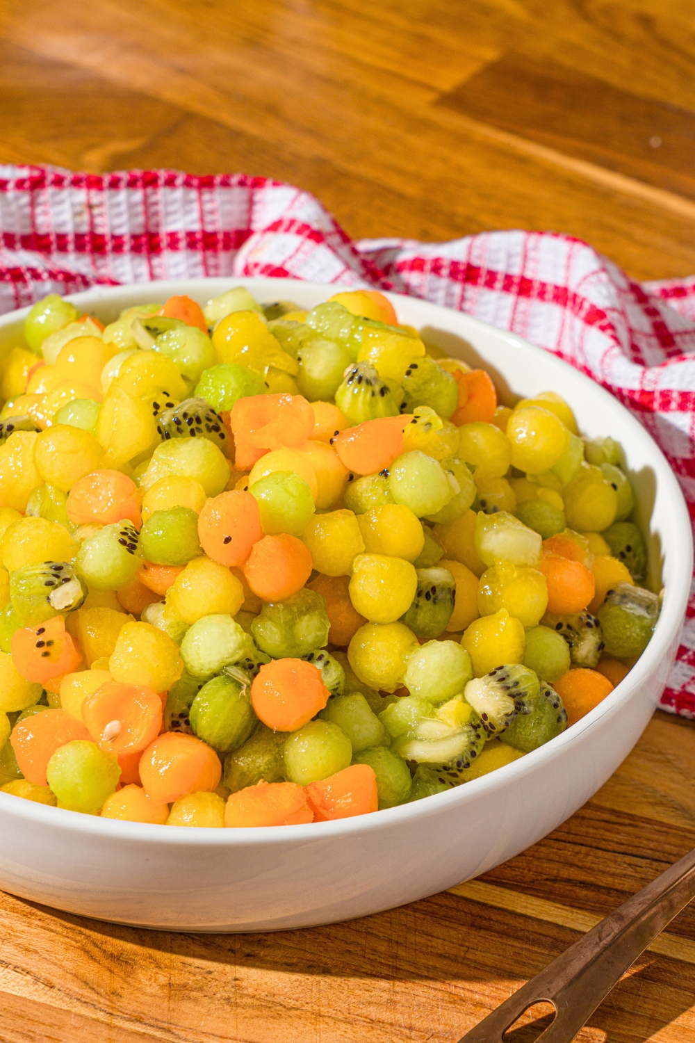 A dot fruit salad in a white. The fruit salad is small balls of yellow watermelon, honey dew, kiwi, and apricots.