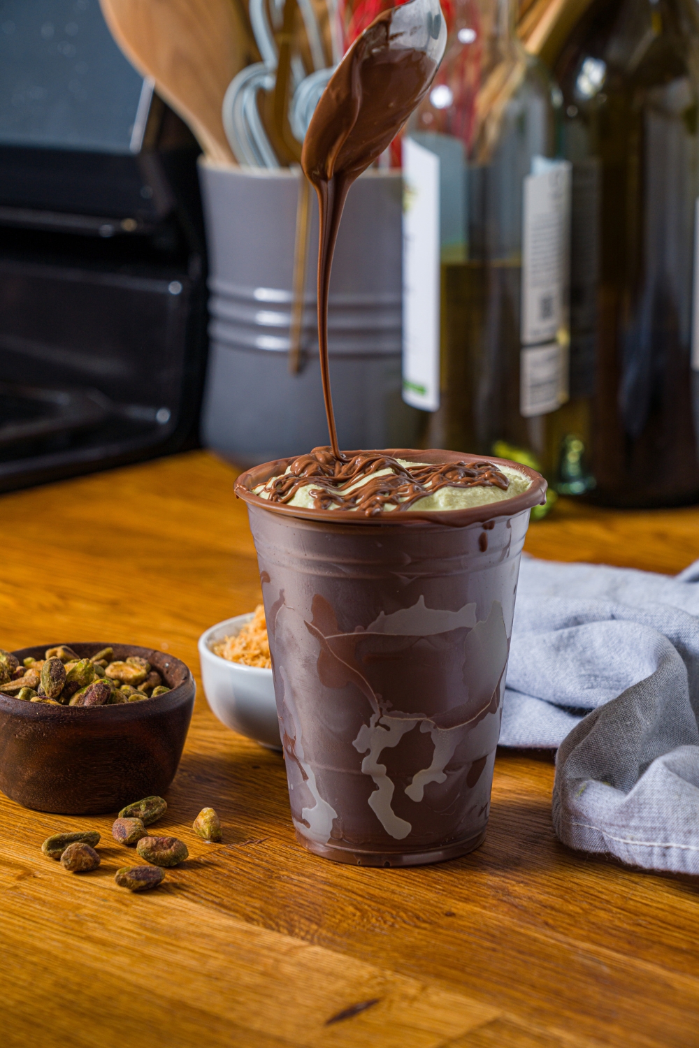 A spoon drizzling chocolate on top of a Dubai chocolate milkshake in a plastic cup that is lined completely with chocolate.