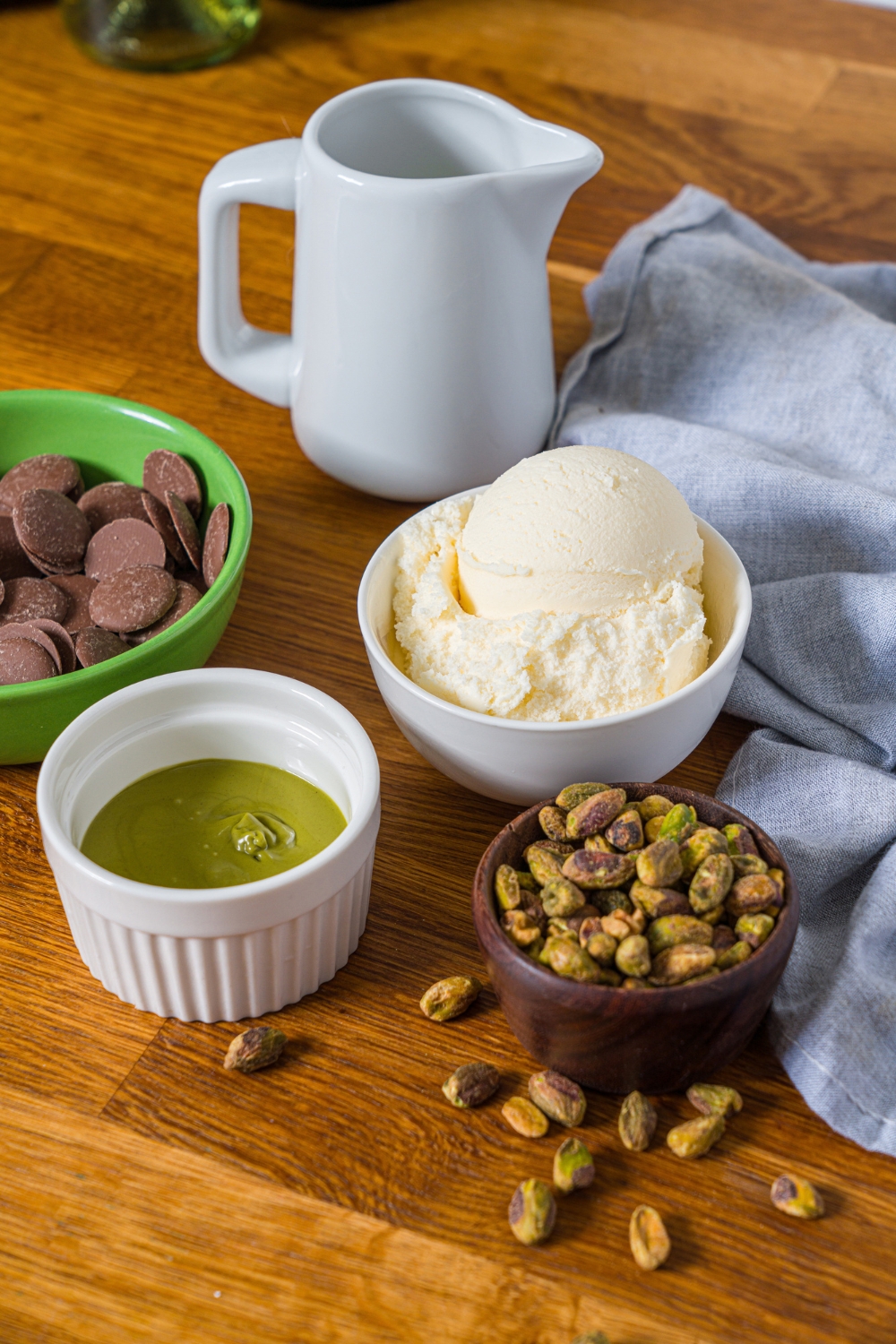 A bowl of vanilla ice cream, part of a green cup with chocolate melting wafers in it, a white bowl of pistachio cream, and a bowl of pistachios all on a wooden counter.