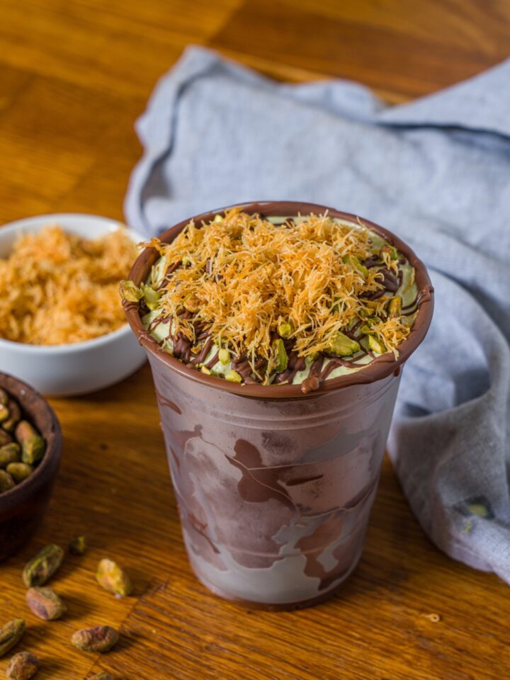 A Dubai chocolate shake in a chocolate lined plastic cup. On top of the shake is drizzled, chocolate, pistachios, and katafi.