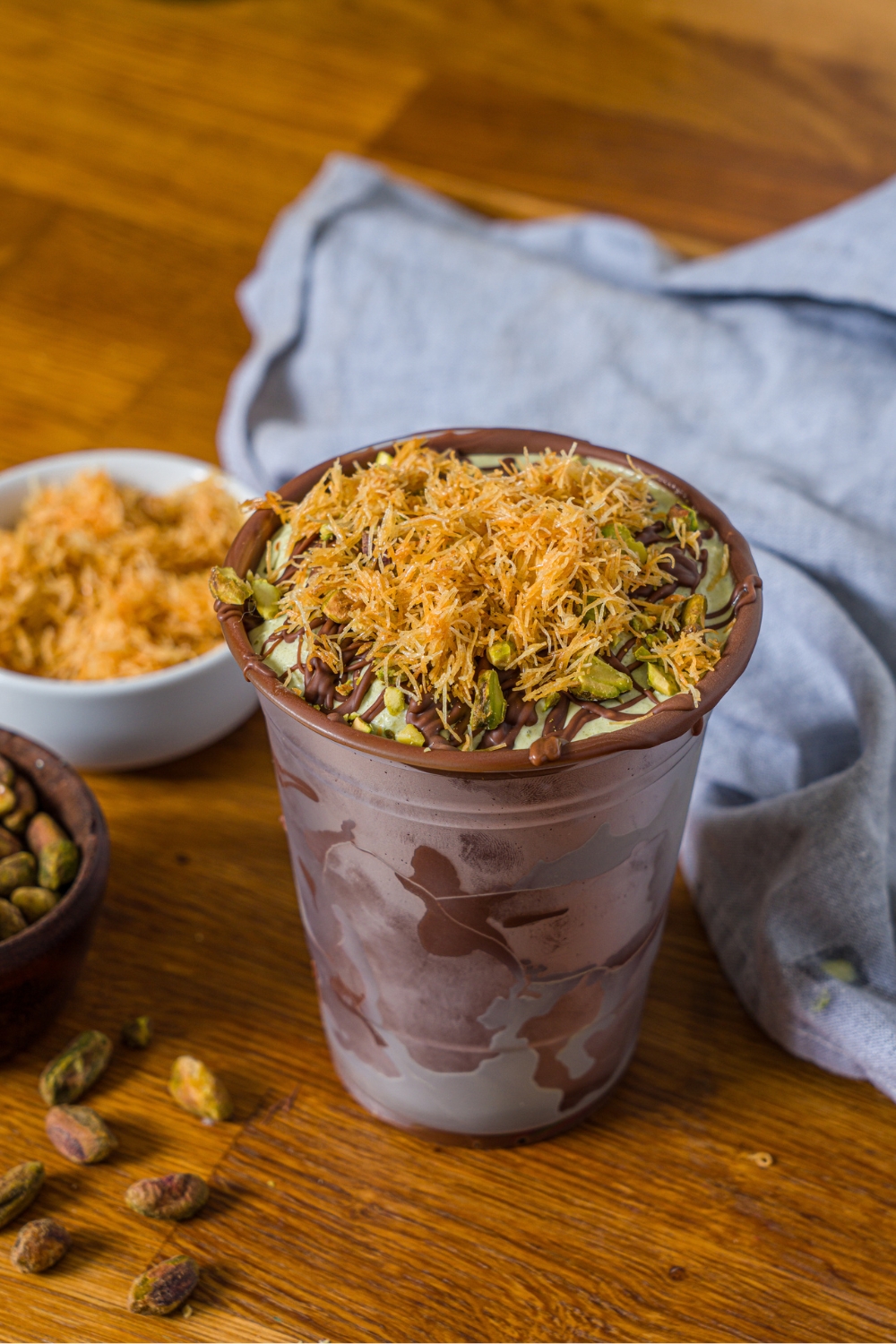 A Dubai chocolate shake in a chocolate lined plastic cup. On top of the shake is drizzled, chocolate, pistachios, and katafi.