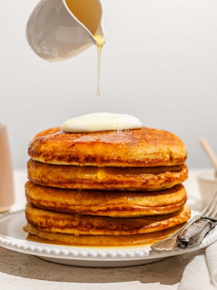 A pitcher, pouring honey butter, on top of a stack of four golden diner pancakes on a white plate.