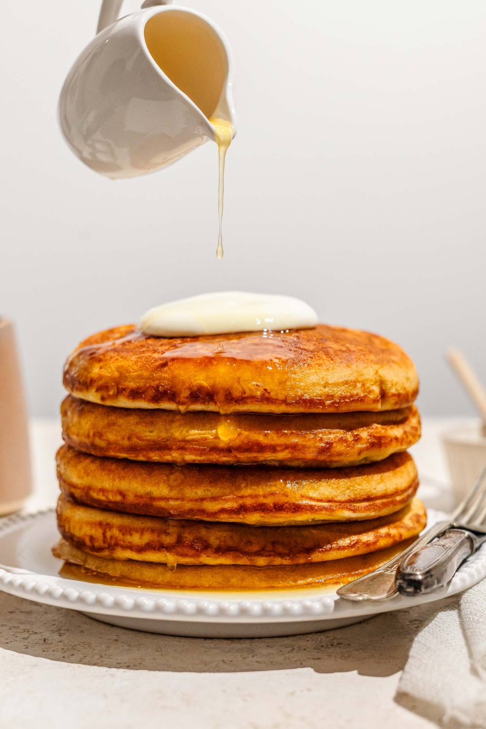 A pitcher, pouring honey butter, on top of a stack of four golden diner pancakes on a white plate.
