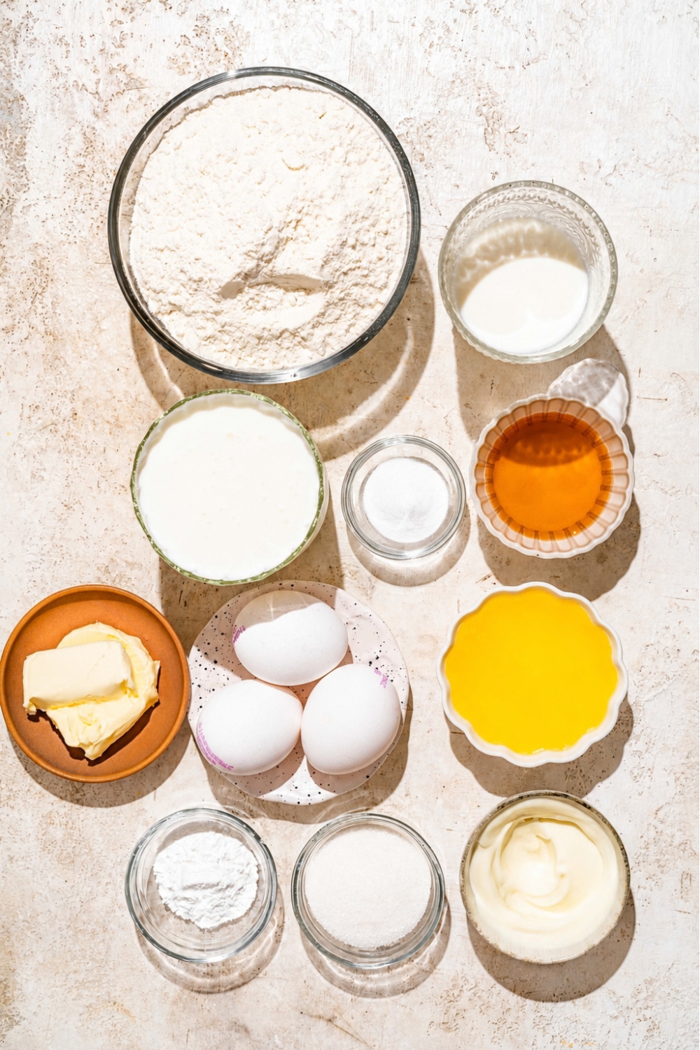 Golden diner, pancakes ingredients, including a bowl of flour, a bowl, buttermilk, a bowl of baking powder, three eggs, cup of butter, a cup of melted butter, a cup of honey, a cup of sugar, all on a white counter.