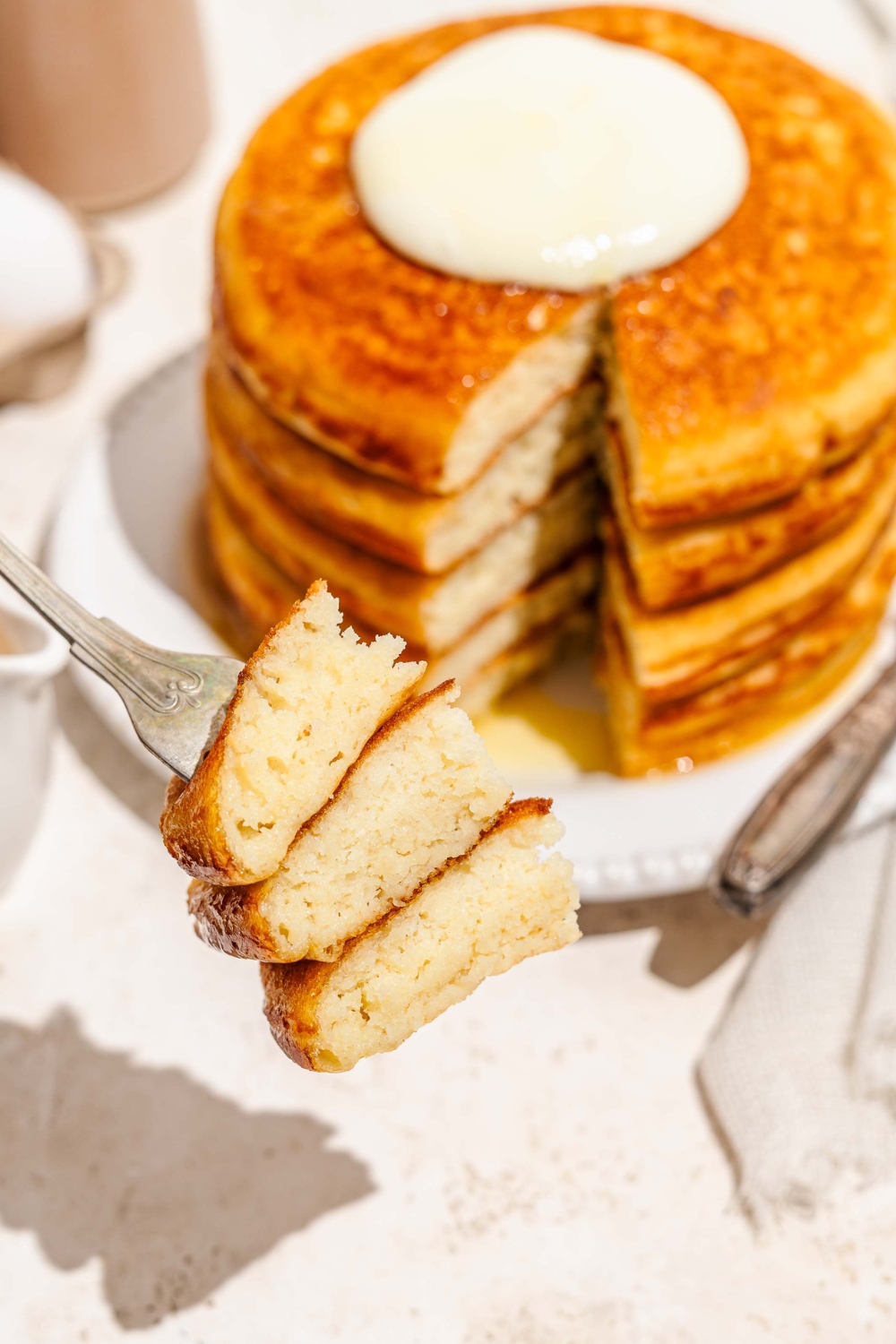 A fork that has three triangle slices of fluffy golden diner pancakes on it. The fork is holding it in front of a stack of the pancakes with a slice missing from it.