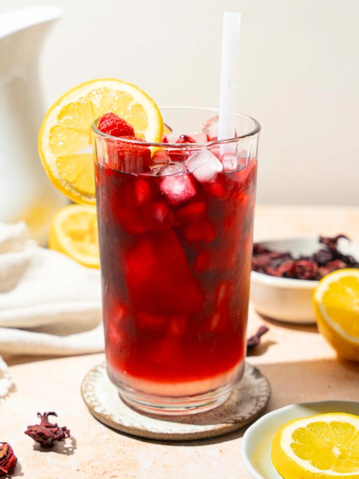 A tall glass of hibiscus lemonade served with ice, sliced lemon, and a straw. The glass is on a coaster on a tan counter with a white cloth napkin, sliced lemon, and a bowl of hibiscus tea leaves.