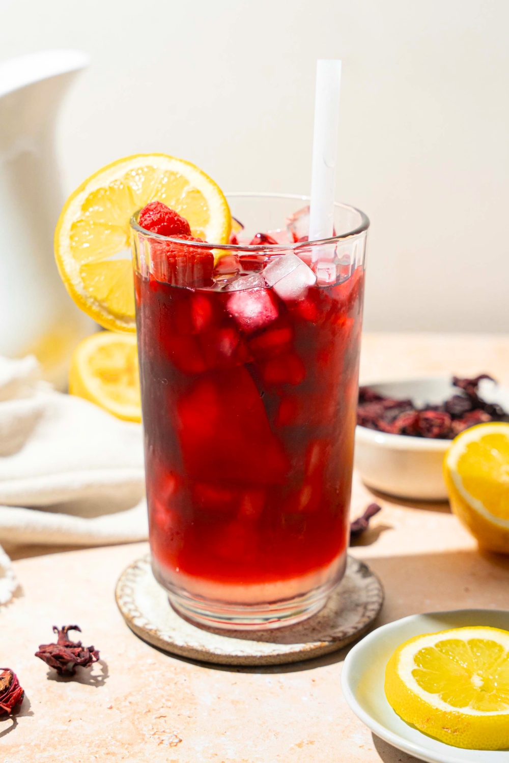 A tall glass of hibiscus lemonade served with ice, sliced lemon, and a straw. The glass is on a coaster on a tan counter with a white cloth napkin, sliced lemon, and a bowl of hibiscus tea leaves.
